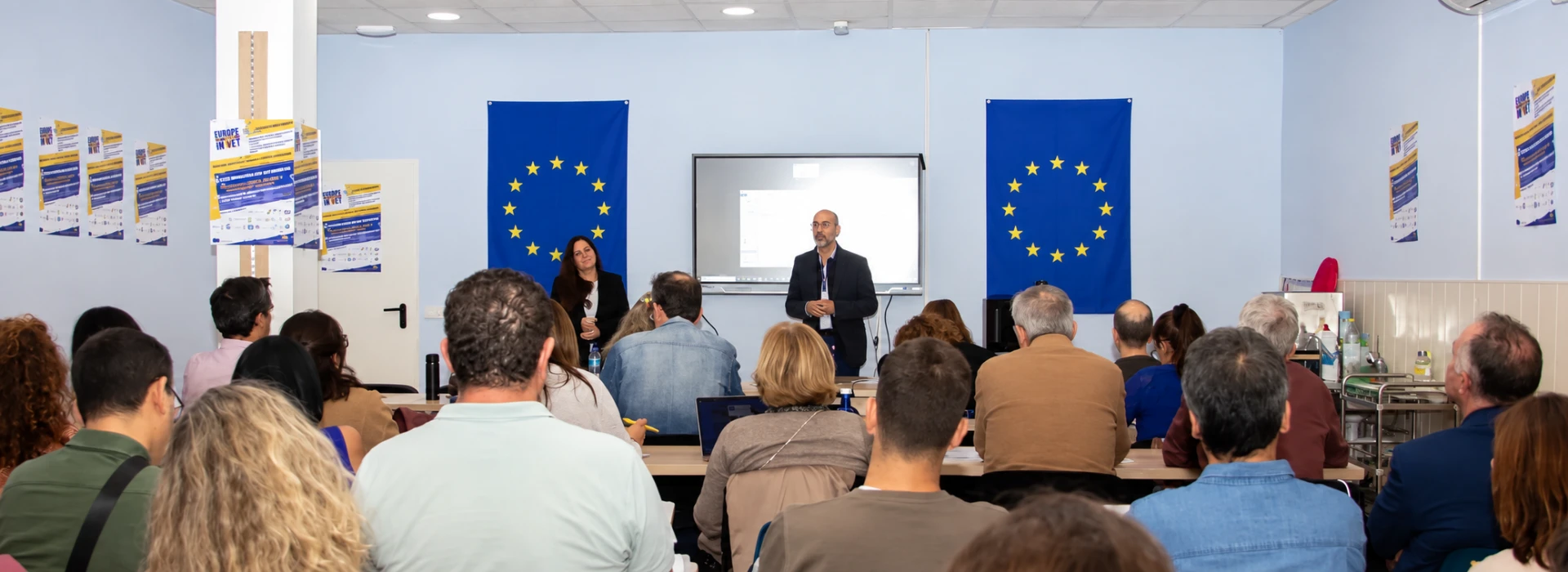 Two speakers addressing education-centre representatives between European Union flags at the 2025 consortium meeting